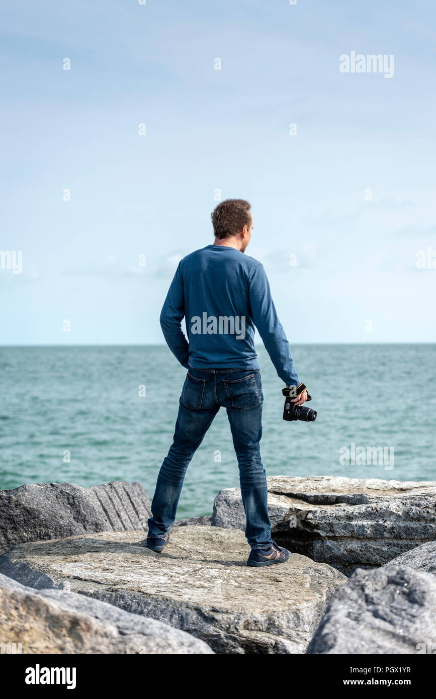 Homme debout sur des rochers surplombant la mer tenant un appareil photo, vue de dos. Banque D'Images Homme debout sur des rochers surplombant la mer tenant un appareil photo, vue de dos. Banque D'Images