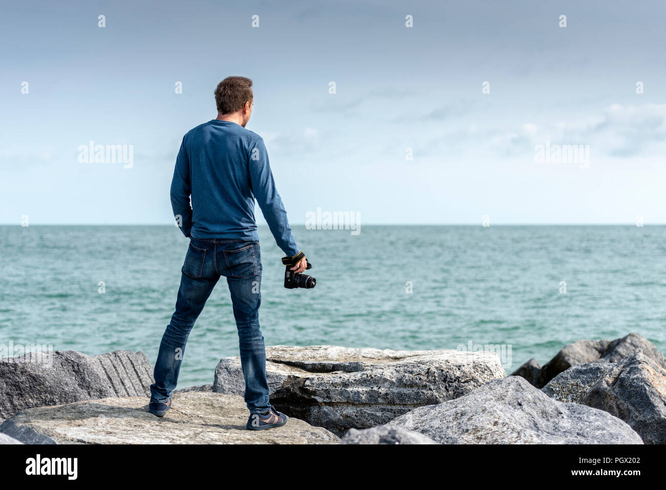 Homme debout sur des rochers surplombant la mer tenant un appareil photo, vue de dos. Banque D'Images Homme debout sur des rochers surplombant la mer tenant un appareil photo, vue de dos. Banque D'Images