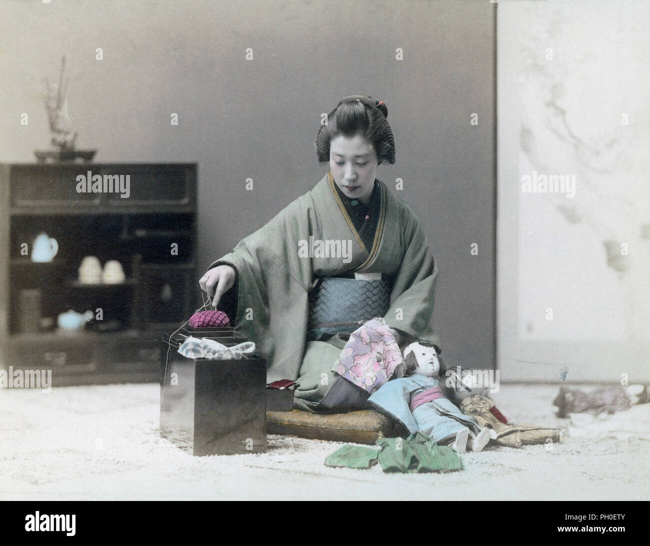 [ 1890 - Japon femme japonaise en Kimono Couture ] - une jeune japonaise en kimono traditionnel et casquettes Vêtements pour poupées couture est assis sur un zabuton coussin. Deux poupées s'appuyé contre ses genoux. Un kit de couture avec un pin cushion se tient sur le sol en face d'elle. 19e siècle vintage albumen photo. Banque D'Images