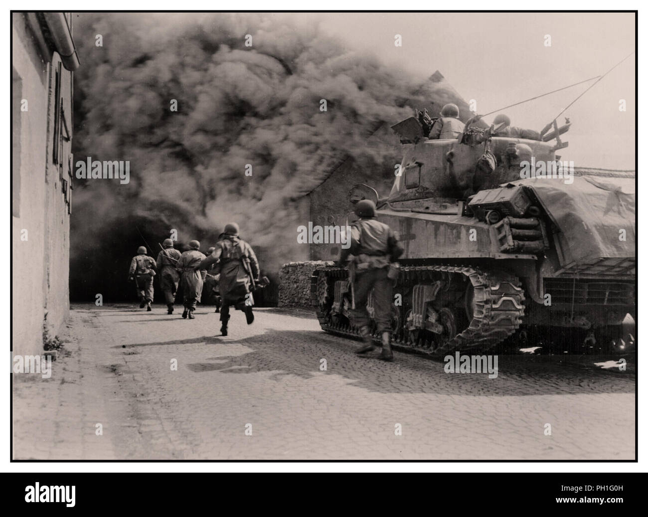 WW2 GI soldats du 55e Bataillon d'infanterie blindée et armée troisième char Sherman M4 du 22e bataillon de chars, l'avance se déplacer à travers la fumée remplie street. Wernberg, Allemagne. 22 avril 1945 Banque D'Images