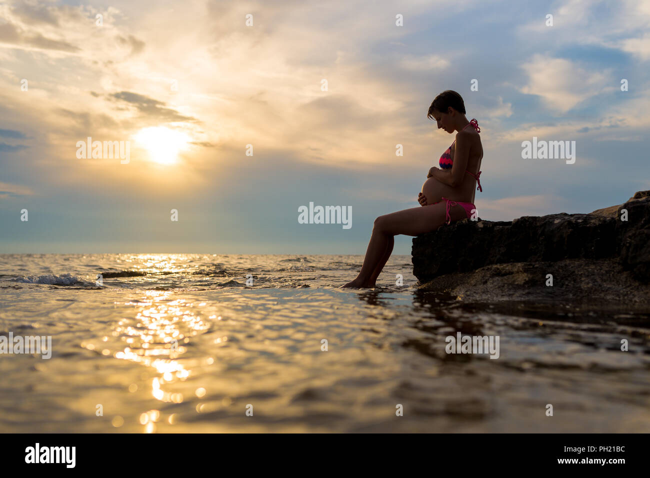 Femme enceinte en maillot assis de profil, berçant son ventre sur un rocher au bord de la mer au coucher du soleil avec le soleil jette une golden beam partout e Banque D'Images