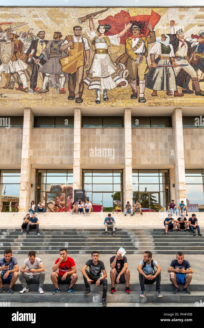 Les jeunes se détendre sur les étapes de la Musée Historique National, ci-dessous, l'ère communiste, mosaïque murale, dans la place Skanderbeg, Tirana, Albanie, Banque D'Images