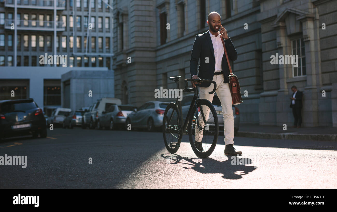 Jeune homme d'affaires la marche à l'extérieur avec un vélo et à l'aide de téléphone mobile. Homme africain d'aller travailler en vélo de conversations au téléphone cellulaire sur rue. Banque D'Images