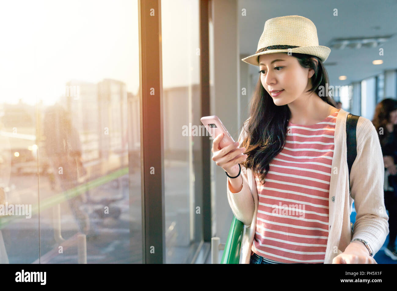 Belle asiatique femme parcourt son smartphone debout à côté de grandes fenêtres. Banque D'Images