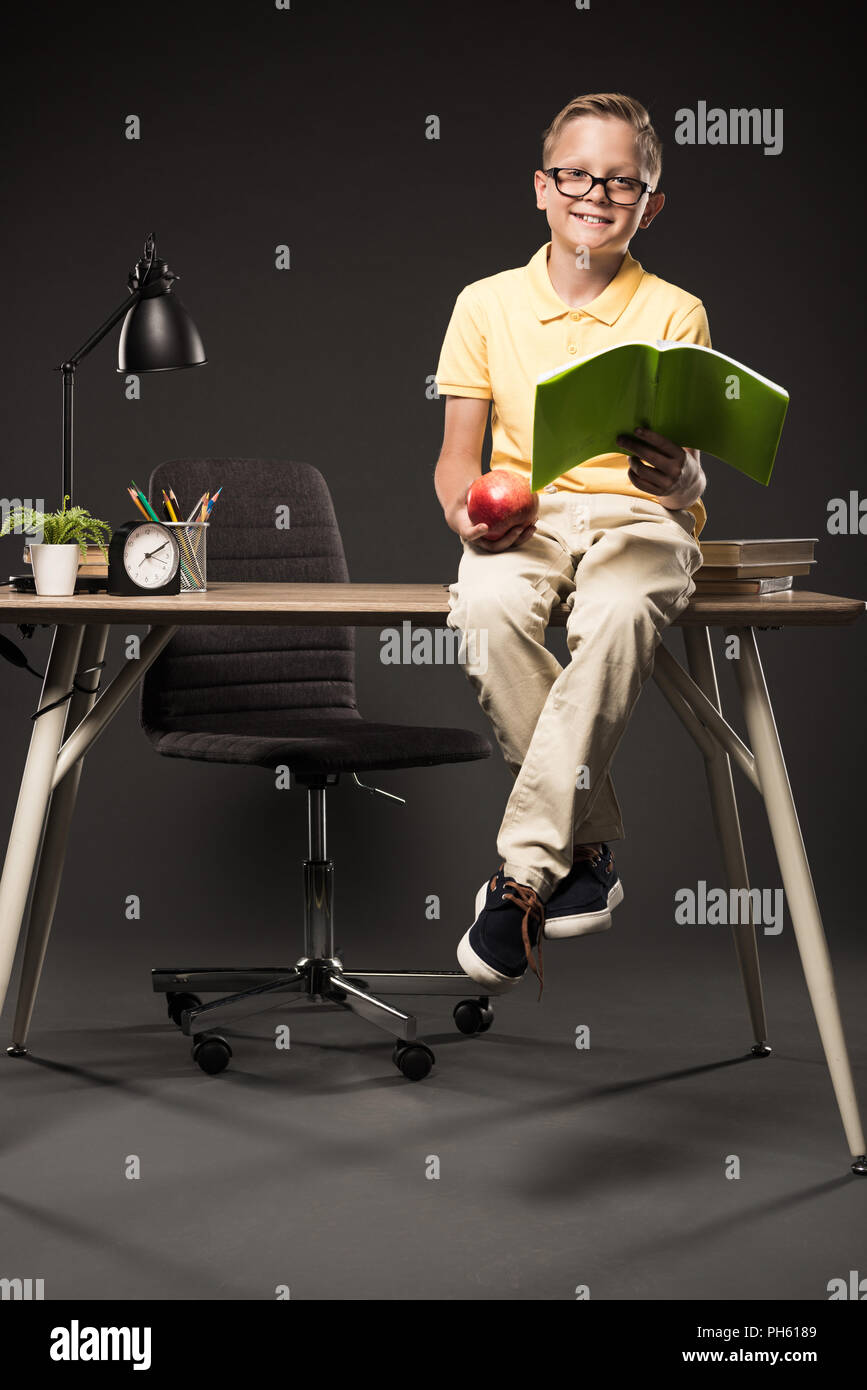 Smiling businesswoman holding apple et faire leurs devoirs tout en étant assis sur la table avec des livres, des plantes, lampe, crayons de couleur, réveil et manuel sur gray backgrou Banque D'Images