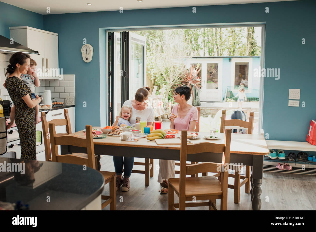 Vue grand angle de trois mères adultes assis à une table de cuisine à parler. Deux des mères ont leurs enfants avec eux et profiter de certains nac Banque D'Images