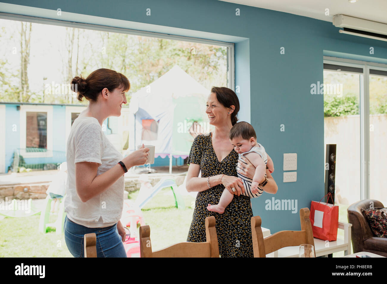 Deux femmes adultes debout dans la socialisation et à la maison possèdent. Une femme tient son bébé. Ils sont parler et rire ensemble. Banque D'Images