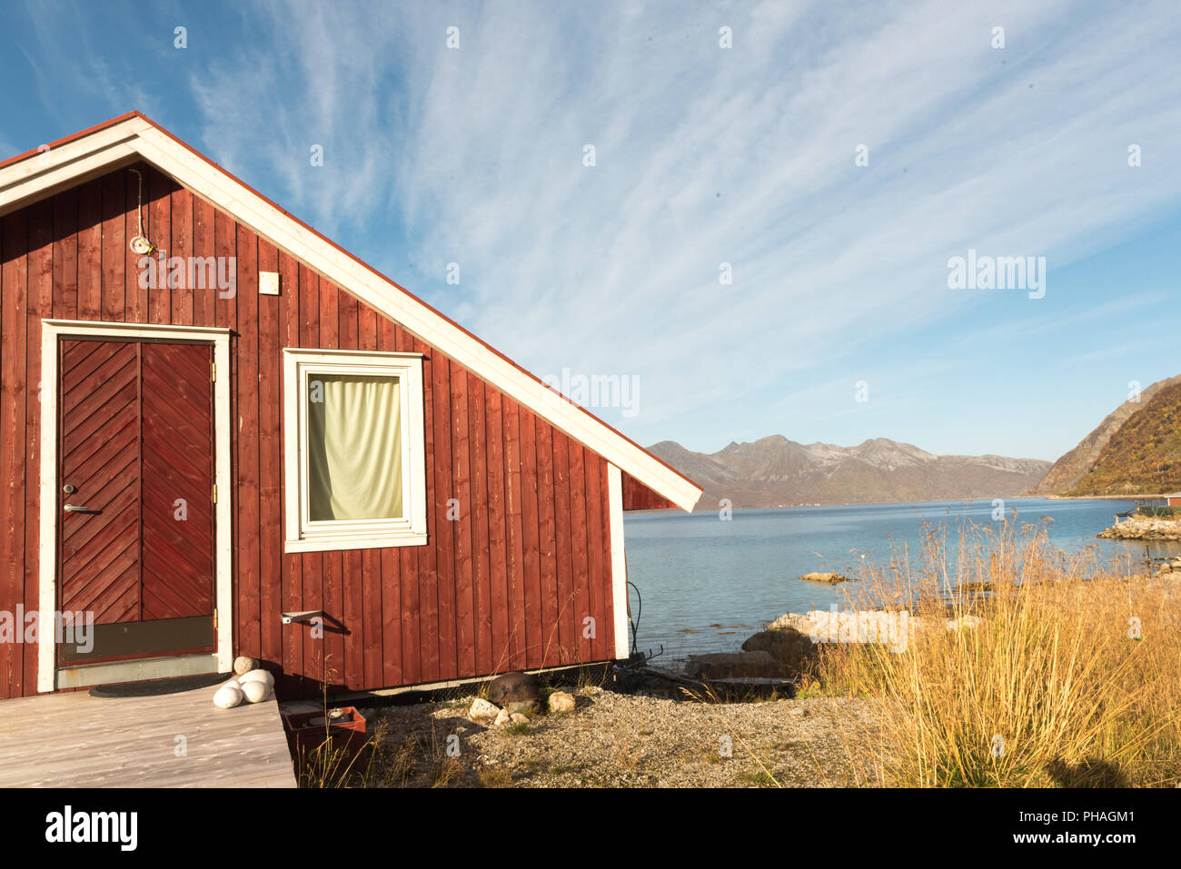 Vue d'un Norvégien en bois cabane de plage avec des pierres sur le rivage avec vue sur le Fjord de Tromso, Norvège et Cercle arctique, sommet de montagne sous le soleil bleu Banque D'Images