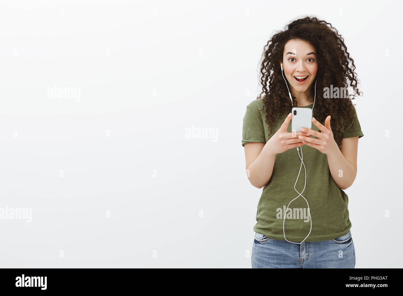 Aucun moyen, son incroyable. Portrait d'impressionné et surpris charming brunette avec des cheveux bouclés, laissant tomber la mâchoire et le regard rêveur à huis clos, à l'écoute de la musique dans les écouteurs et holding smartphone Banque D'Images