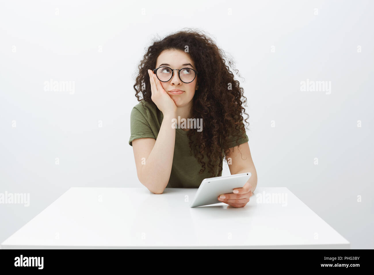 Portrait d'imprudents de rêve belle étudiante à lunettes noires, regardant vers le haut et se penchant sur le visage tandis que la main assis à table, holding digital smartphone, l'ennui et l'indifférence Banque D'Images