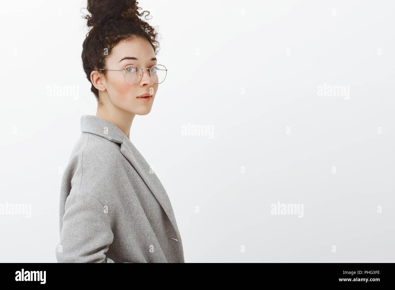 Jusqu'à la taille de balle sûr smart girl élégante avec des cheveux bouclés peignées à bun, debout dans le profil et dans les verres à l'appareil photo, être féminine et tendre à l'arrière-plan gris Banque D'Images