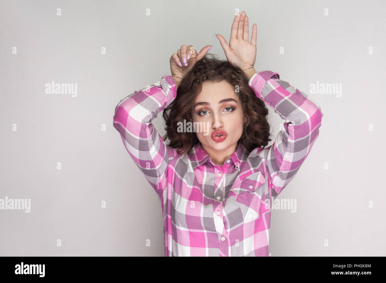 Belle fille drôle avec chemise à carreaux roses, curly hairstyle et un miroir et permanent à la caméra avec bunny à mains geste. Piscine studio shot Banque D'Images