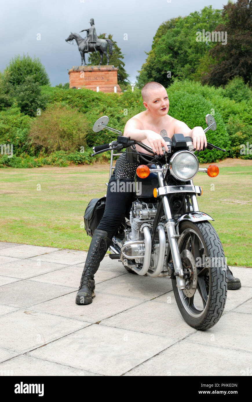 Rosa Fury assis sur une moto Suzuki, ayant fait signe . Elle porte une  courte robe noire et collants résille et dispose d'un casque de moto en  face d'elle Photo Stock -
