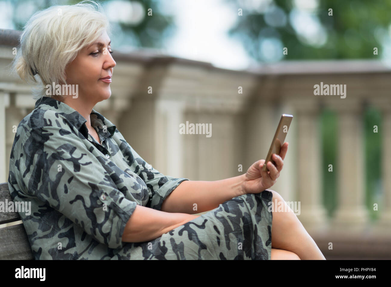 Jolie femme assise sur un banc extérieur lecture sur son téléphone portable avec un sourire tranquille de plaisir dans un close up side view Banque D'Images