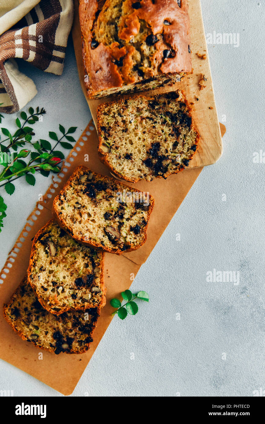 Pain aux courgettes au chocolat avec des noix en tranches sur une planche à découper en bois et placés sur du papier marron accompagné de feuilles vertes sur fond gris Banque D'Images