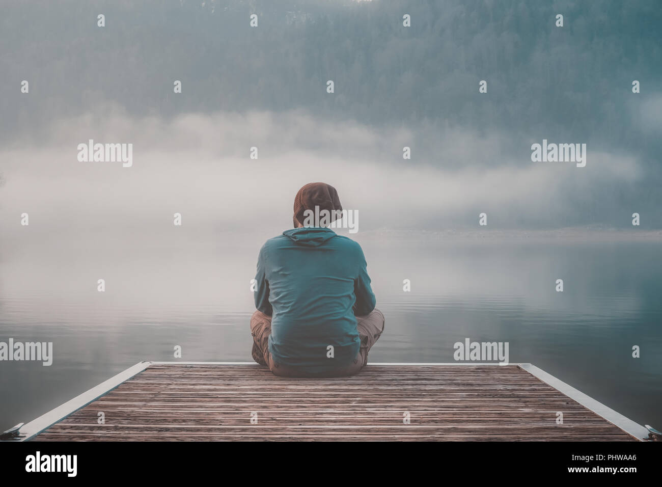 Jeune homme avec capuche assis sur la jetée d'un lac en un jour brumeux. Banque D'Images