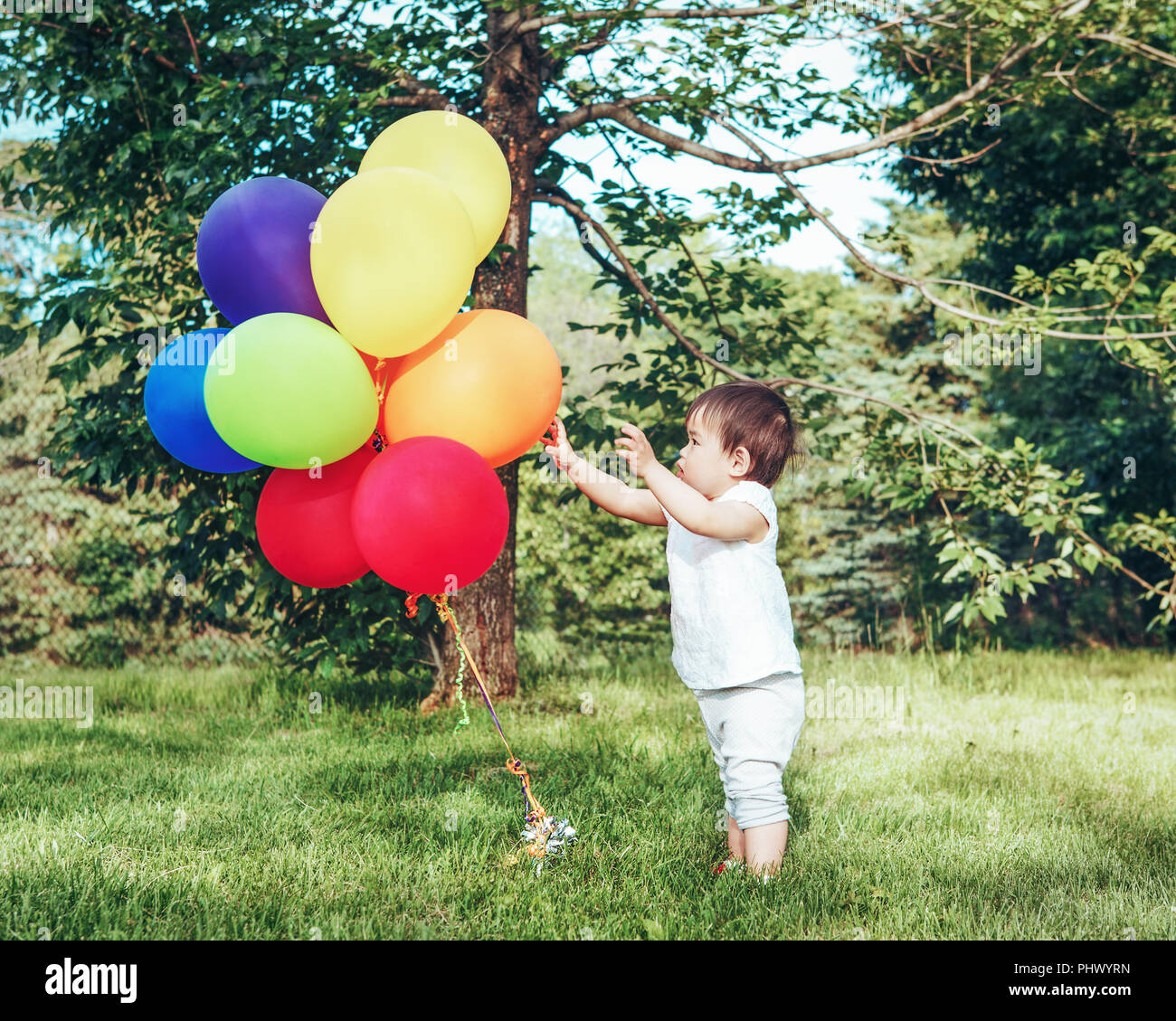 Portrait of cute adorable petite fille asiatique, un enfant de deux ans, en pantalon et chemise blanche, debout avec des ballons sur terrain éclairé forêt prairie Banque D'Images