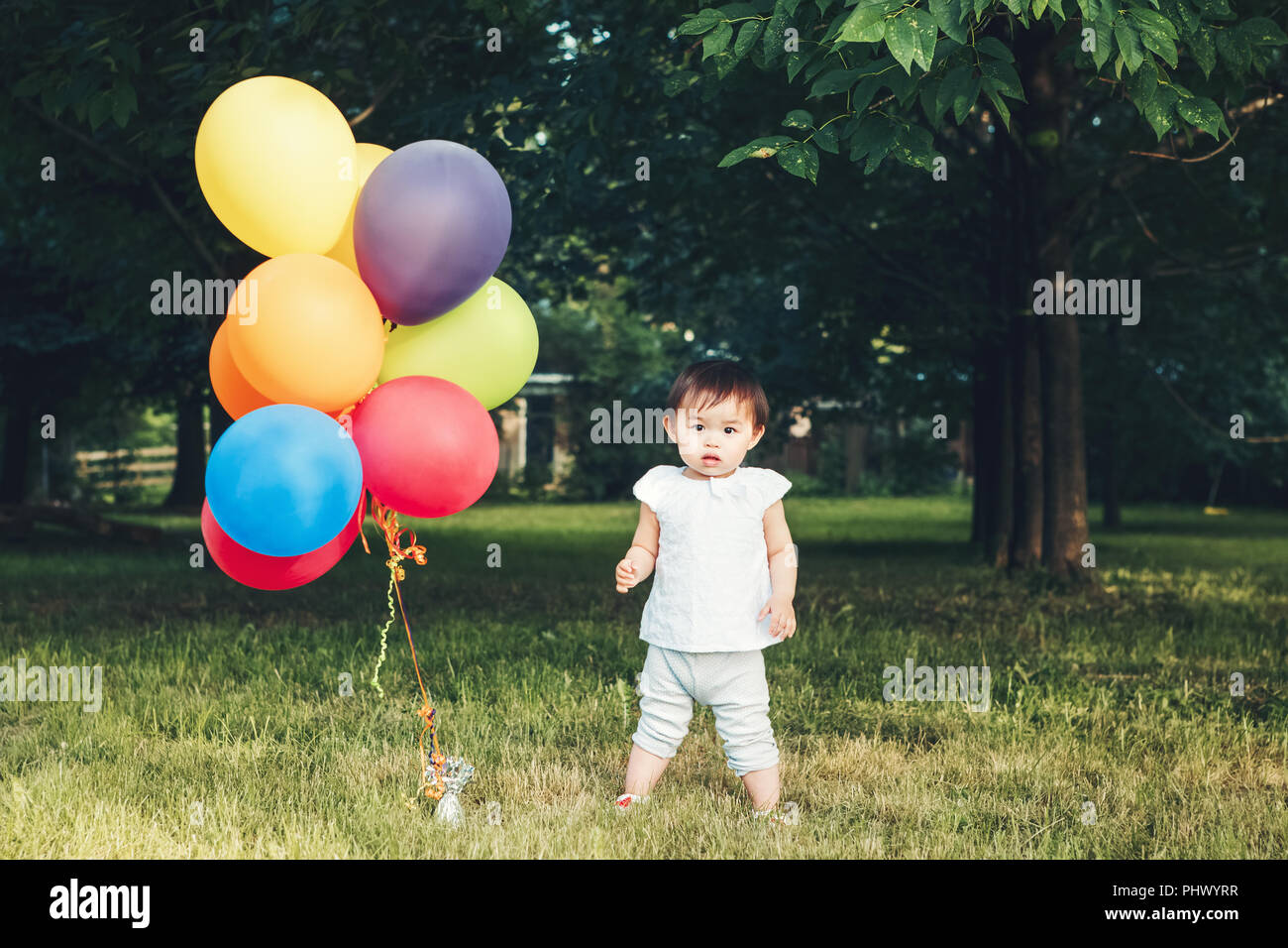 Portrait of cute adorable petite fille asiatique, un enfant de deux ans, en pantalon et chemise blanche, debout avec des ballons sur terrain pré sur le coucher du soleil, happ Banque D'Images