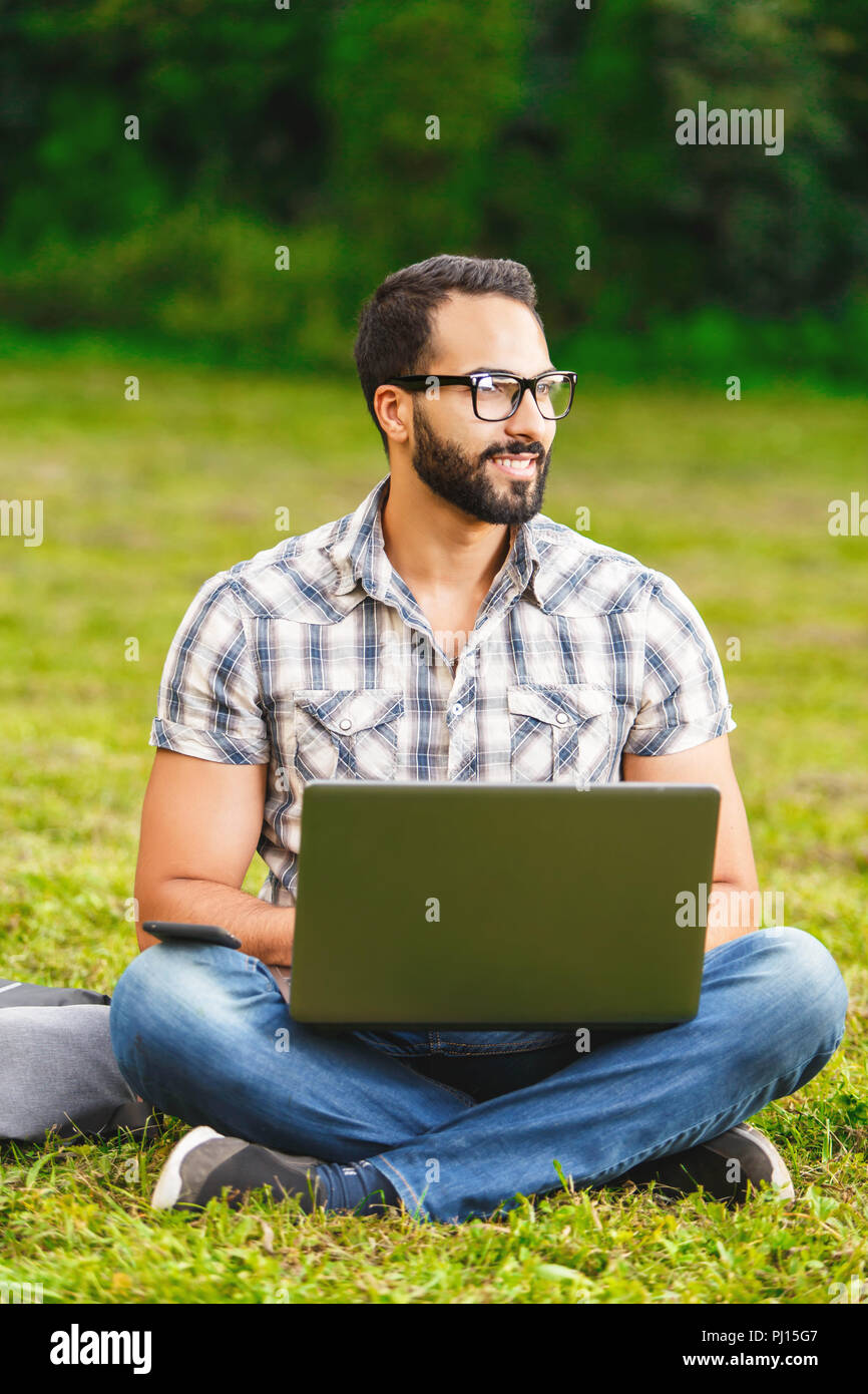 Jeune homme barbu en chemise à carreaux portant des lunettes assis sur l'herbe dans le parc avec son ordinateur portable et à la recherche autour de la belle journée ensoleillée. Banque D'Images