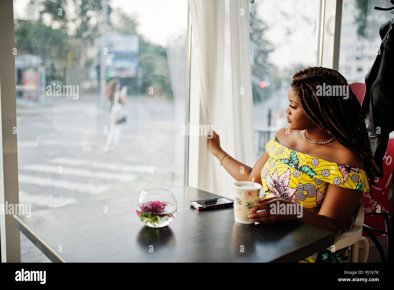 Hauteur petit mignon african american girl avec des dreadlocks, d'usure au niveau de la couleur jaune robe, assis au café avec tasse de café et en regardant la fenêtre. Banque D'Images