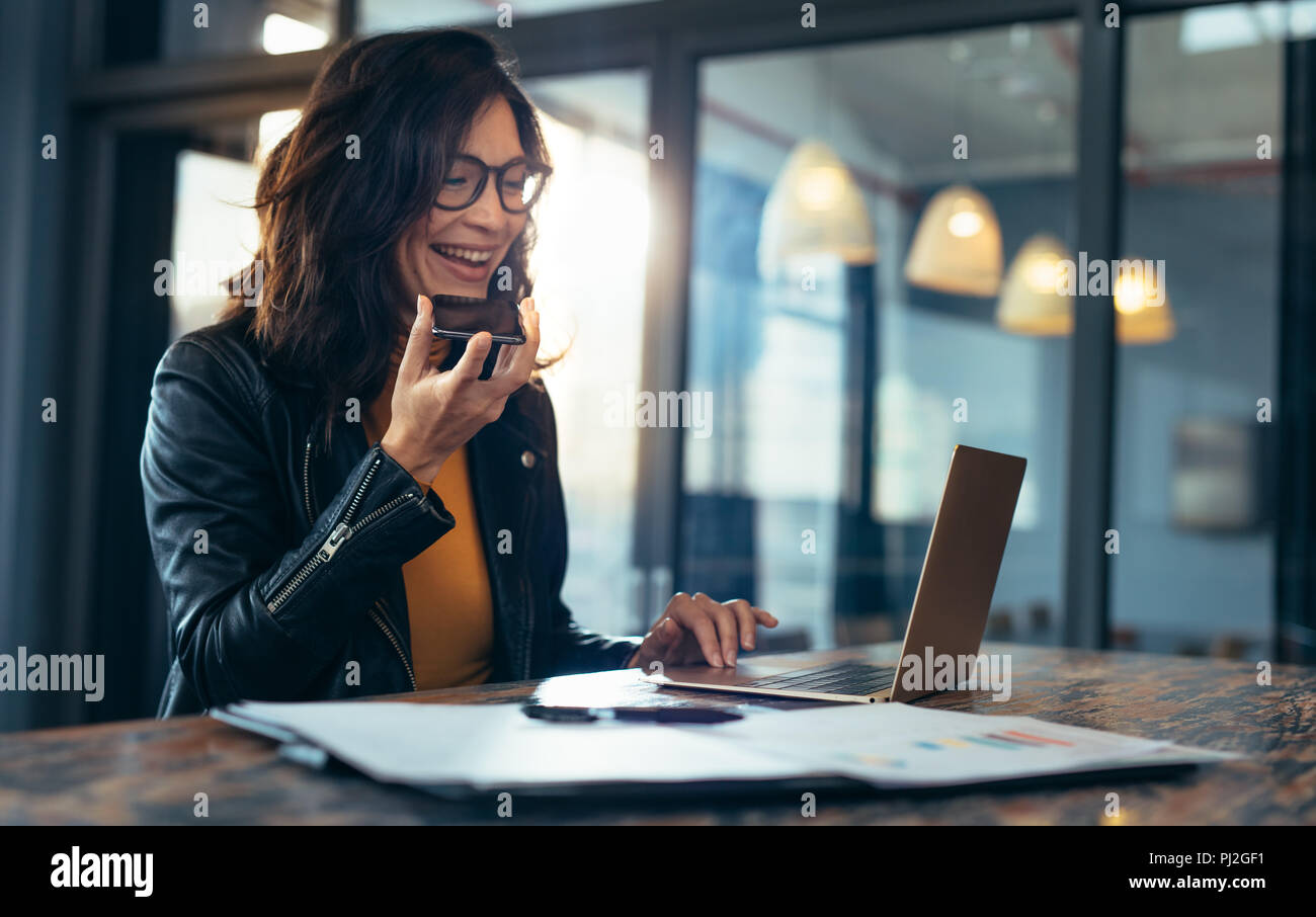 Happy business woman sitting at table et travailler sur ordinateur portable. Femme d'origine asiatique assis au bureau de communication téléphonique ayant une conversation avec Banque D'Images Happy business woman sitting at table et travailler sur ordinateur portable. Femme d'origine asiatique assis au bureau de communication téléphonique ayant une conversation avec Banque D'Images
