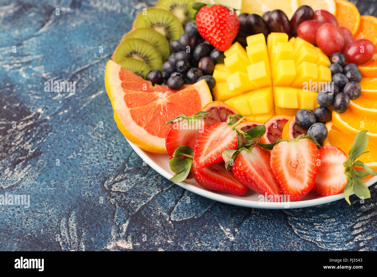Assiette de fruits et baies sains en couleurs arc-en-ciel close up, fraises, mangue, Raisin, Oranges, kiwis, citrons sur le tableau bleu, copie de l'espace pour texte, selective focus Banque D'Images
