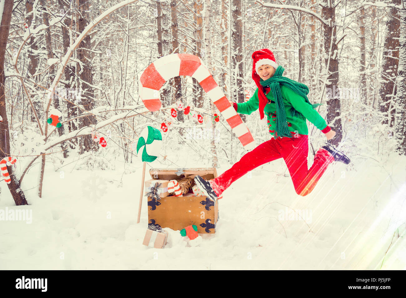 Un homme dans un chapeau de l'an rouge et costume père noël sous gnome dans une gaine verte et rouge pantalon dans la forêt d'hiver avec d'énormes bonbons, une poitrine de cadeaux, le concept de la nouvelle année Banque D'Images