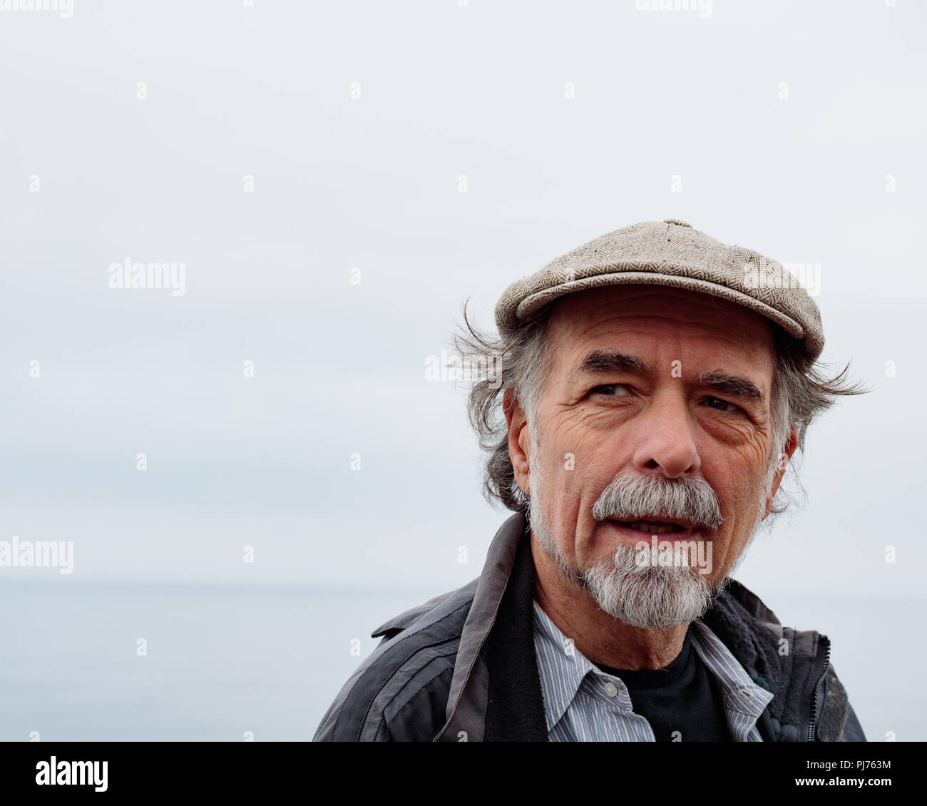 Close up of senior man avec une barbe grise portant une casquette les yeux dans l'horizon de l'océan pâle, distance en arrière-plan Banque D'Images