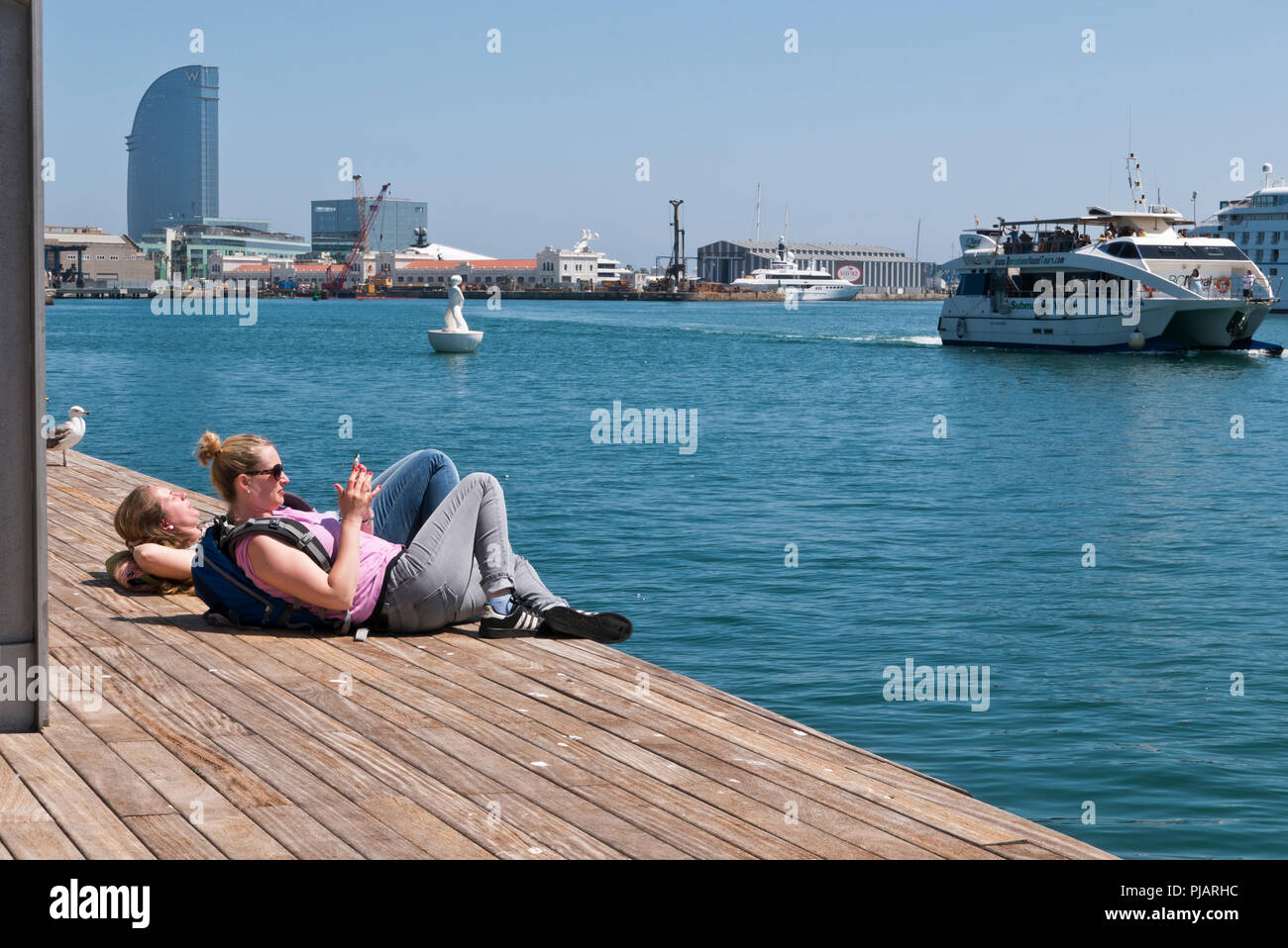 Deux jeunes femmes se trouvant sur le plancher en bois par le bord de la jetée, l'un d'entre eux des messages sur son téléphone portable, Barcelone, Espagne Banque D'Images