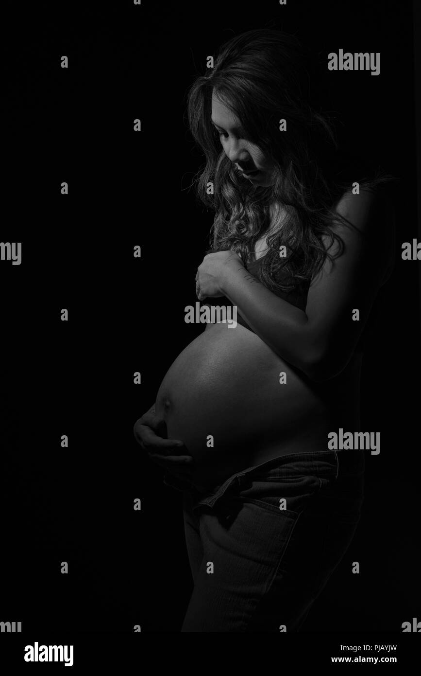 Un pregnant Asian woman posing sur le côté, regardant vers le bas. Un sombre portrait noir et blanc artistique. Banque D'Images