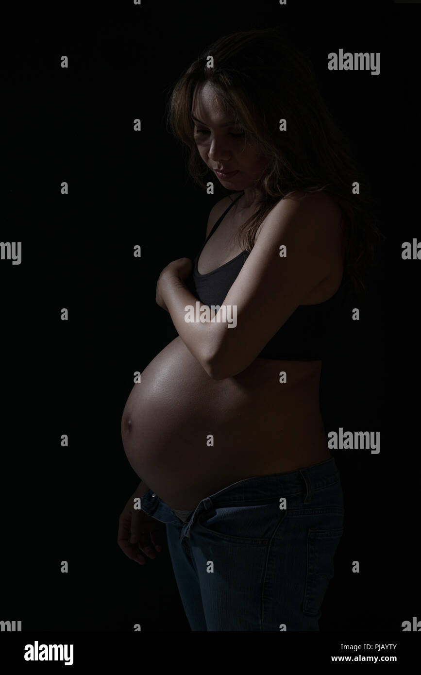 Un pregnant Asian woman posing sur le côté, regardant vers le bas. Un sombre portrait artistique. Banque D'Images