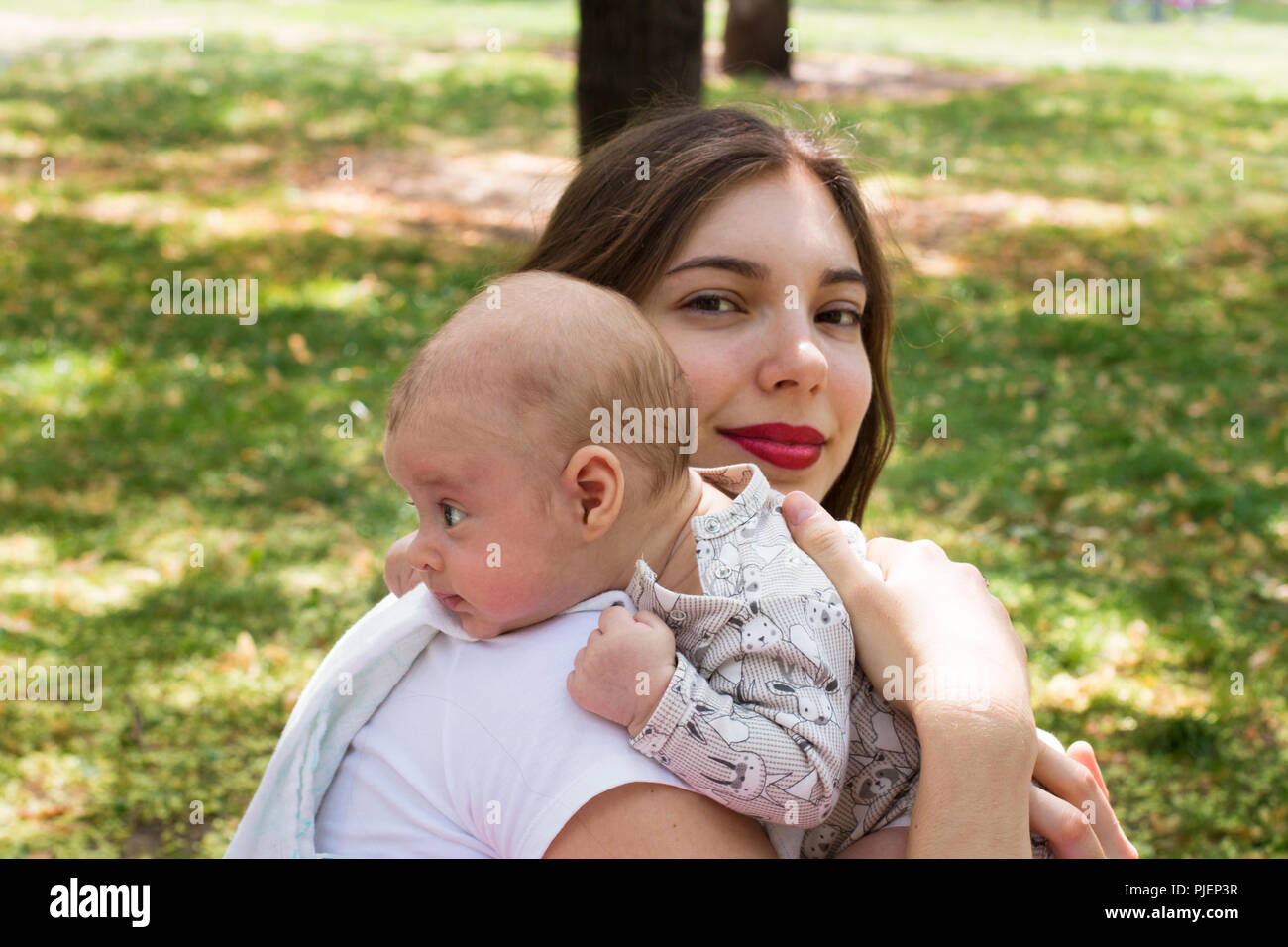 Belle mère tenant son joli bébé à l'extérieur dans le parc. La reposant sur l'épaule de faire un rot en regardant autour, assez heureux maman smiling Banque D'Images