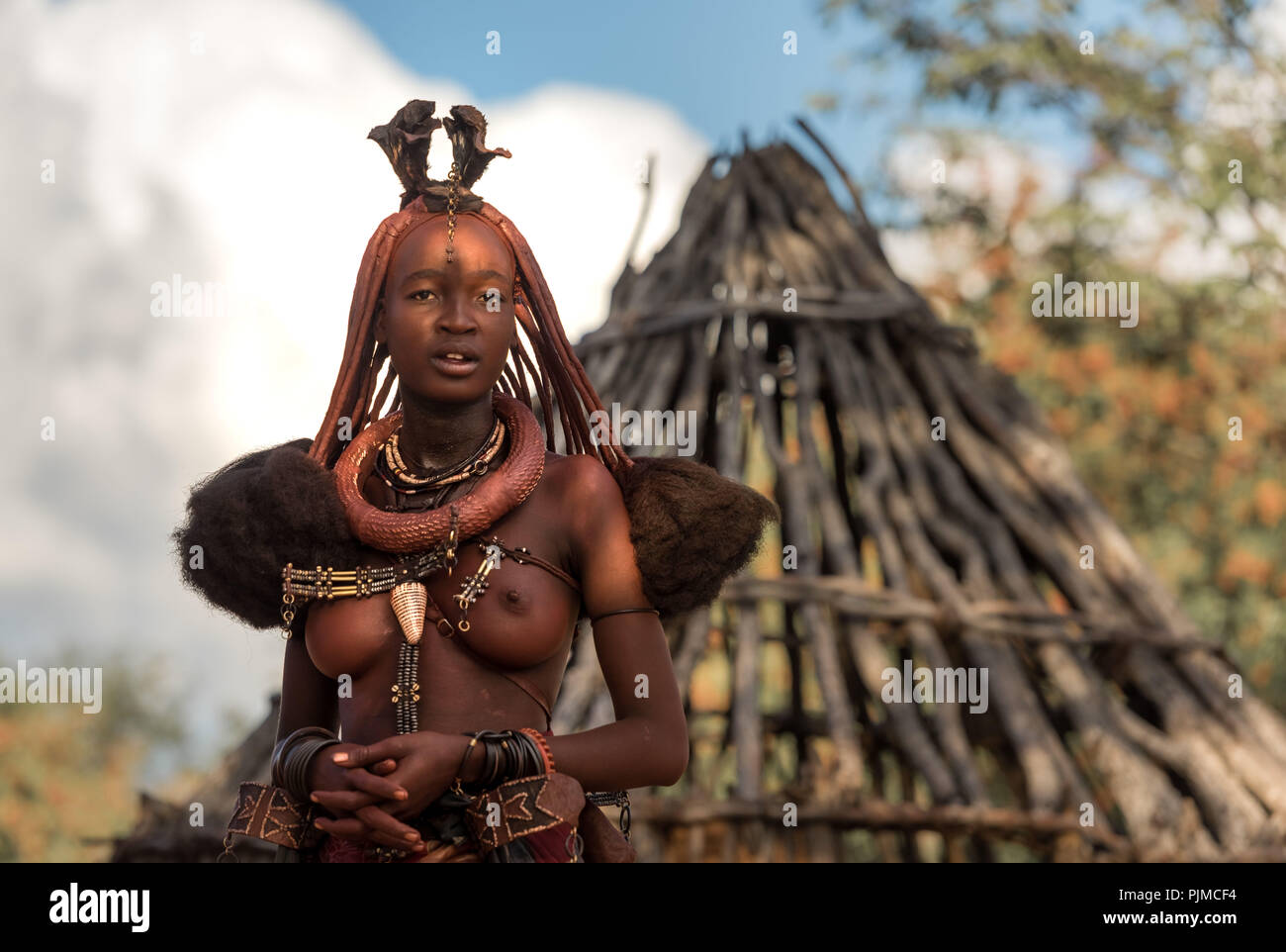 Jolie femme Himba avec de beaux bijoux de corps se trouve dans la mi-ombre dans son village Himba, à la recherche dans l'appareil photo Banque D'Images