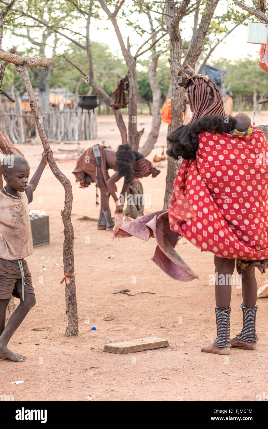 Esprit d'optimisme dans le village Himba : deux femmes Himba recueillir leurs choses ensemble en fin d'après-midi, un garçon Himba est à regarder Banque D'Images