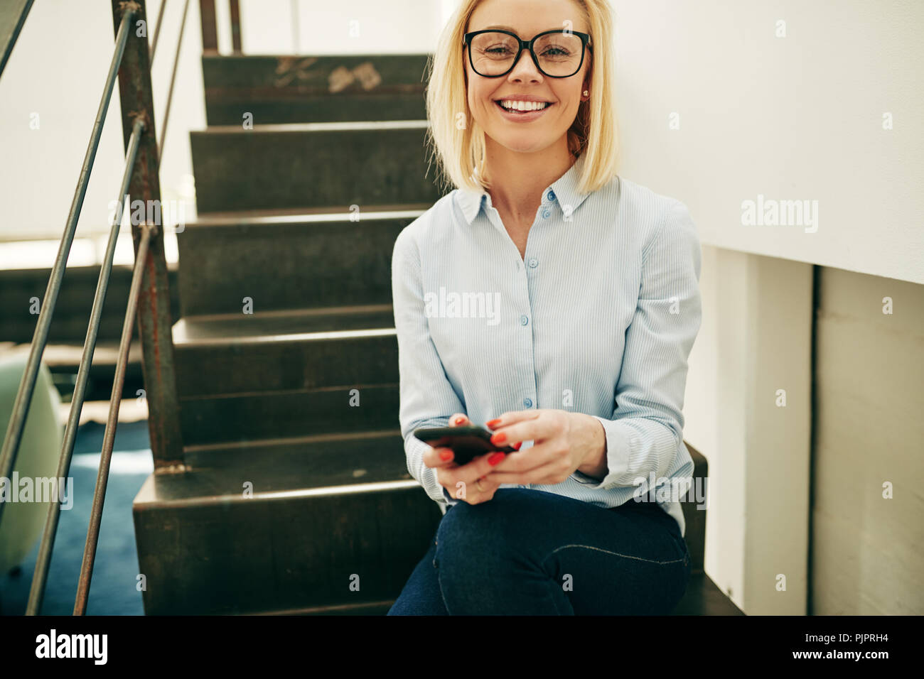 Smiling young businesswoman wearing glasses et la lecture des messages texte sur son portable tout en étant assis dans les escaliers dans un bureau Banque D'Images
