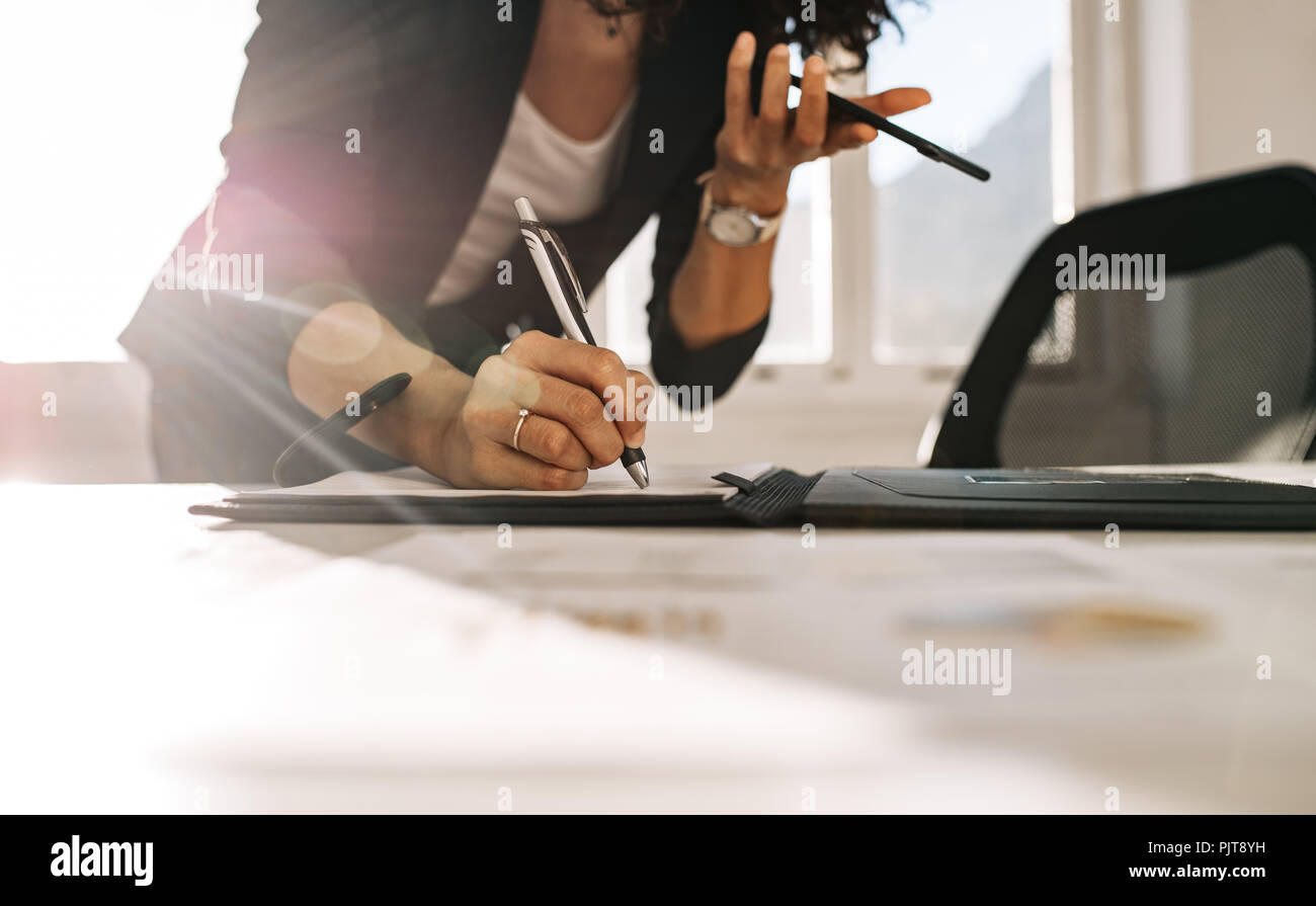 Femme entrepreneur de prendre des notes debout à sa table tout en tenant un téléphone cellulaire dans la main. Close up of a businesswoman writing in journal leani Banque D'Images