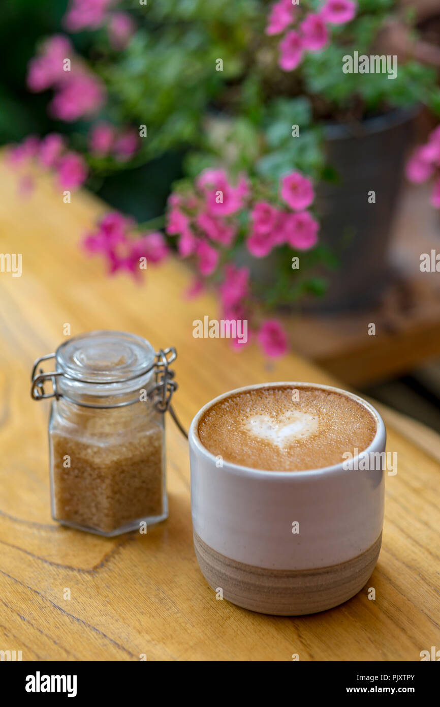 Cardiff, Royaume-Uni. Septembre 2018. Une tasse de café et pot de sucre brun posé sur un banc en bois dans un café avec des fleurs dans l'arrière-plan. Banque D'Images