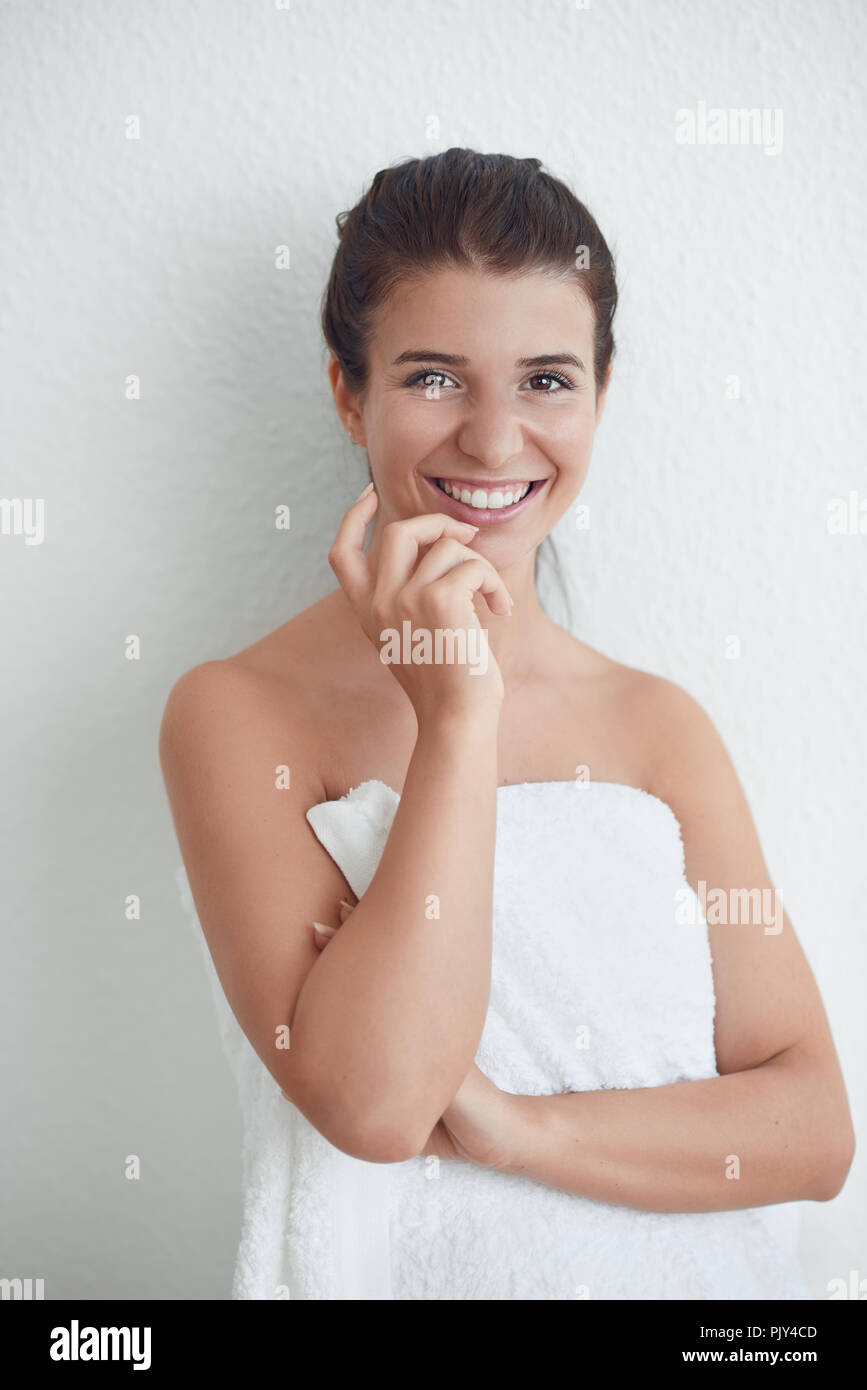 Heureux sain sourire séduisant jeune femme enveloppée dans une serviette blanche pelucheuse propre debout contre un mur blanc de l'hygiène personnelle et conceptuel sp Banque D'Images