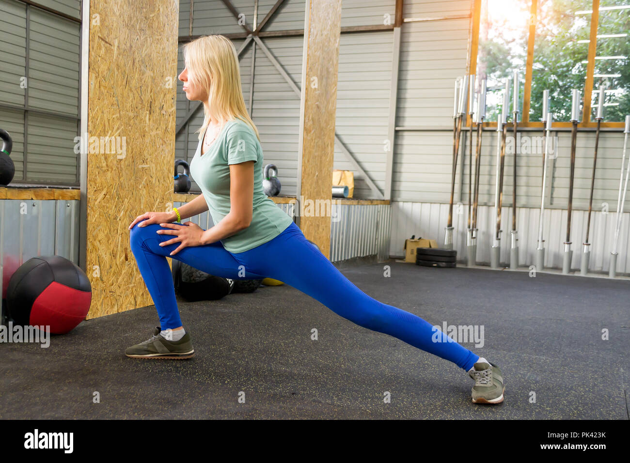 Blonde girl in blue leggins est en train de faire des étirements par fouet  élever sa jambe et appuyé contre le marbre dans la salle de sport sur  l'arrière-plan d'équipement de sport