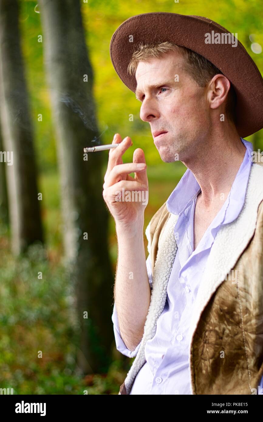 Un homme habillé comme un cow-boy s'appuie contre un arbre fumer une cigarette Banque D'Images