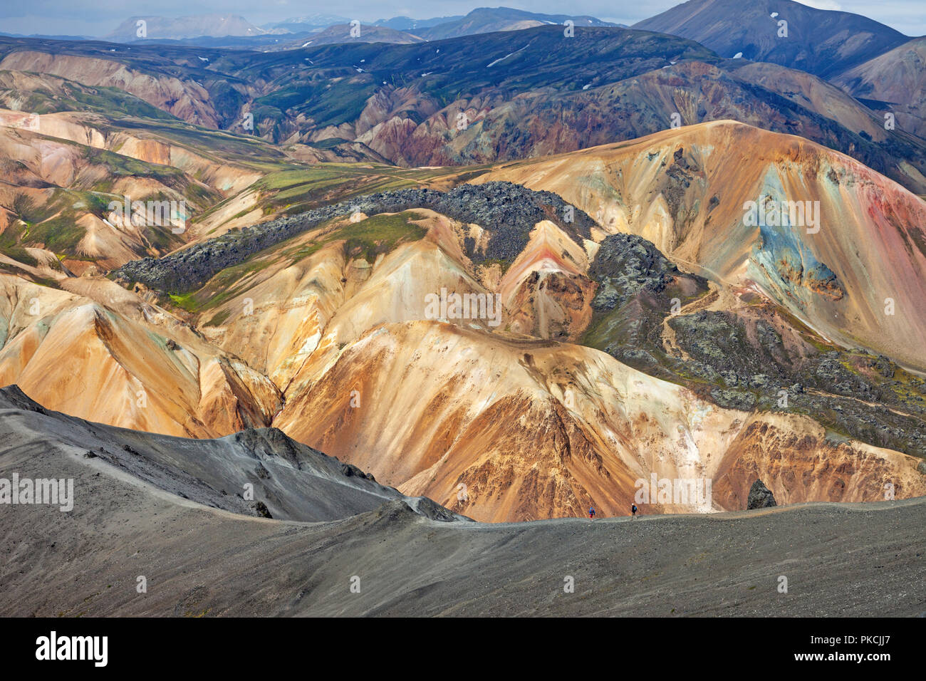 Deux randonneurs descendant la montagne volcanique de Blahnukur avec le Volcan Brennisteinsalda coloré en toile de fond, Landmannalaugar, Islande. Banque D'Images