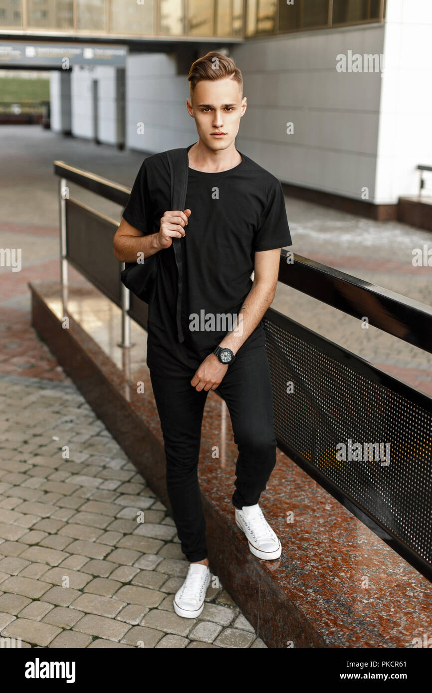 Bel homme élégant hipster en t-shirt noir, pantalon et chaussures blanches  avec un sac à dos, debout près de l'édifice moderne Photo Stock - Alamy
