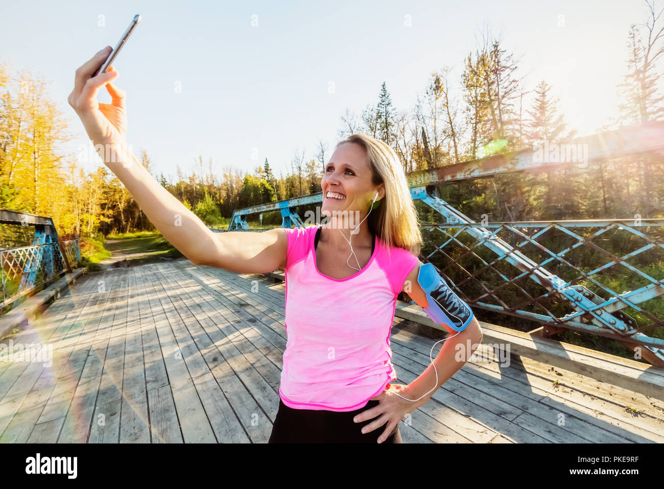 Une femme portant des vêtements et un brassard pour son téléphone portable se tient sur un pont dans un parc en automne en prenant un auto-portrait avec son téléphone portable Banque D'Images