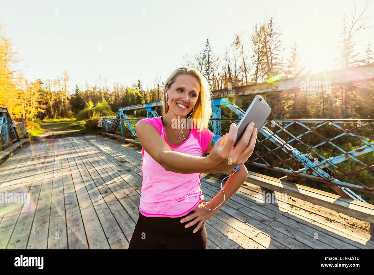 Une femme portant des vêtements et un brassard pour son téléphone portable se tient sur un pont dans un parc en automne en prenant un auto-portrait avec son téléphone portable Banque D'Images