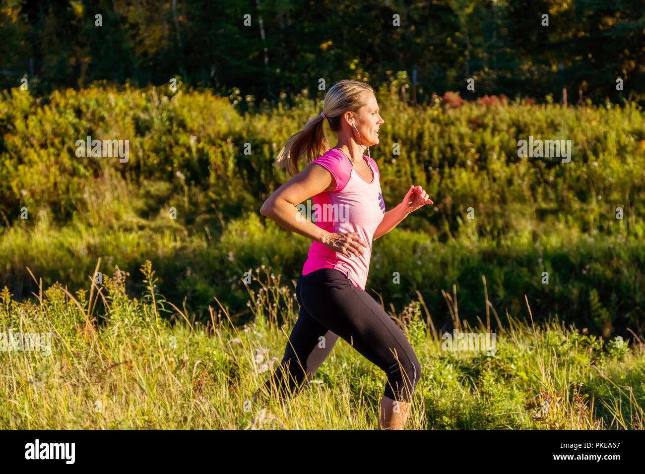 Une belle femme d'âge moyen portant des vêtements et à l'écoute de musique court le long d'un ruisseau dans un parc de la ville au coucher du soleil sur une chaude soirée d'automne Banque D'Images