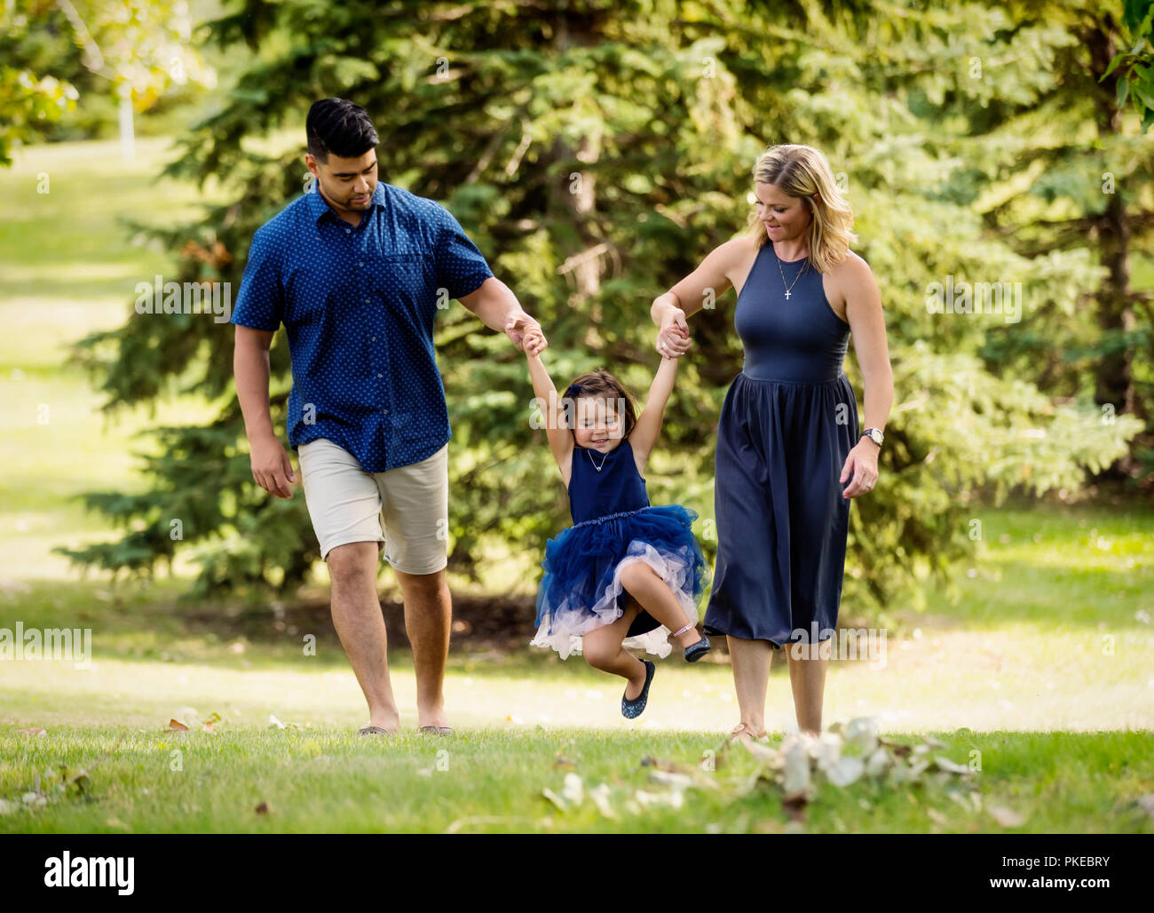 Une mère et son père dans une course à pied mariage mixte et balançant leur petite fille dans l'air lors d'une sortie en famille dans un parc sur une chaude journée d'automne Banque D'Images