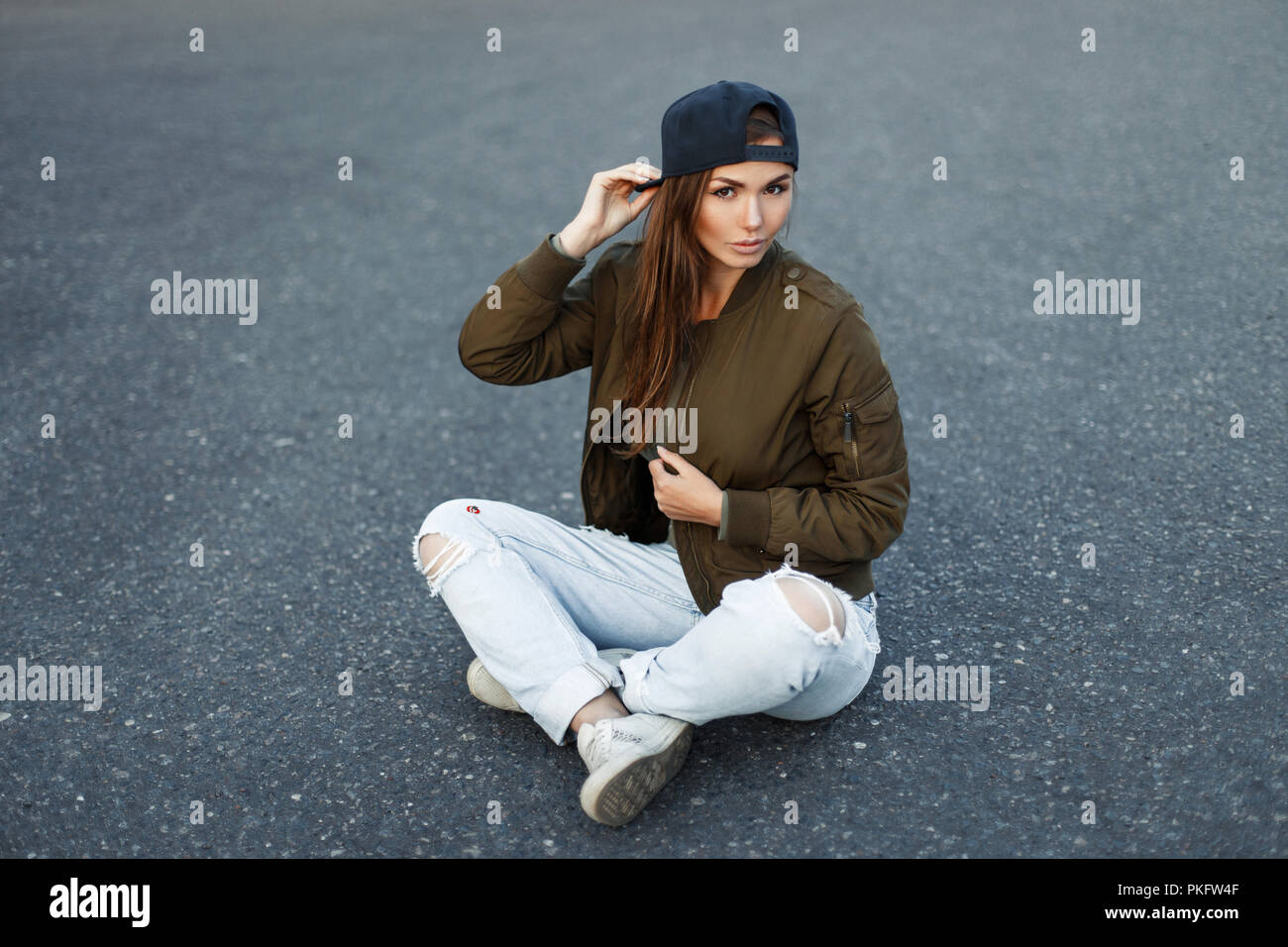 Vogue femme modèle dans une casquette de baseball dans un élégant vert  veste et jeans déchirés est assis sur l'asphalte Photo Stock - Alamy