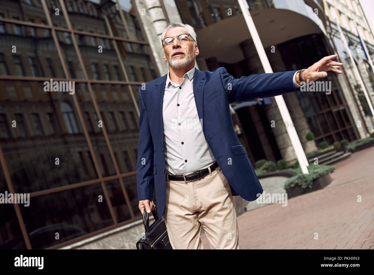 Confiant senior businessman catching taxi en levant son bras et standing outdoors with cityscape dans l'arrière-plan Banque D'Images