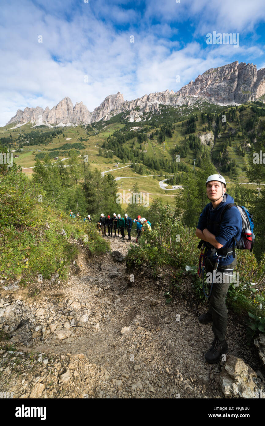 Young male climber regarde une via ferrata dans les Dolomites en Alta Badia avec grand paysage et groupe de grimpeurs derrière lui Banque D'Images