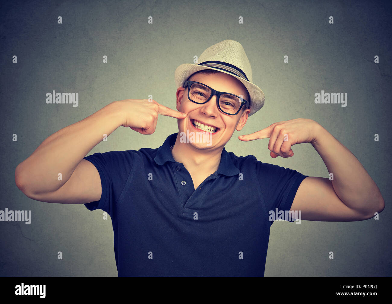 Jeune homme élégant avec chapeau et lunettes en montrant des dents blanches lumineuses donnant sourire à huis clos sur fond gris Banque D'Images